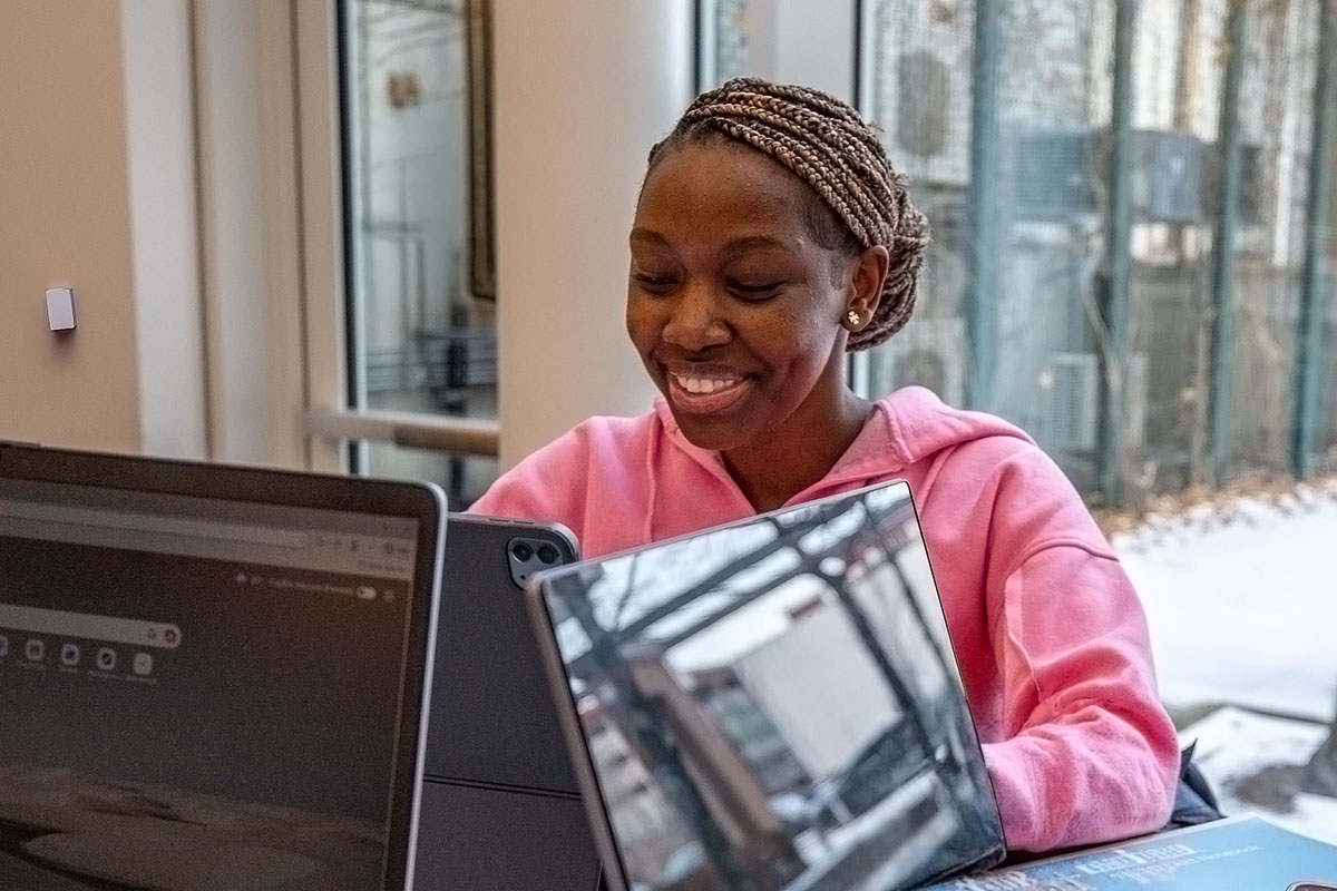 A student, wearing a pink hoodie, smiles while working at a desk with a laptop.