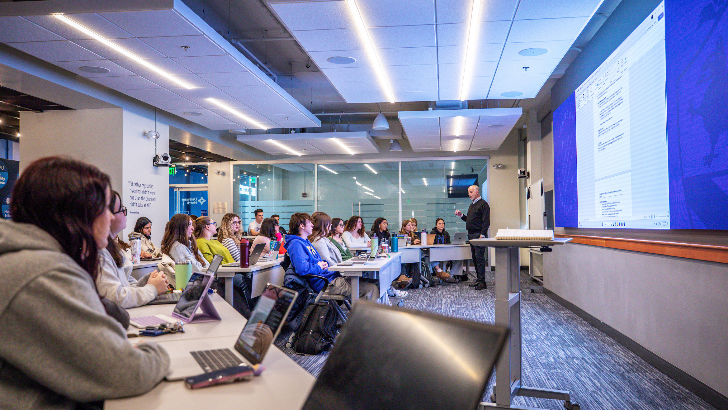 A lecturer stands at a podium in a modern classroom, addressing a group of students seated at desks with laptops, while a large screen displays presentation content.
