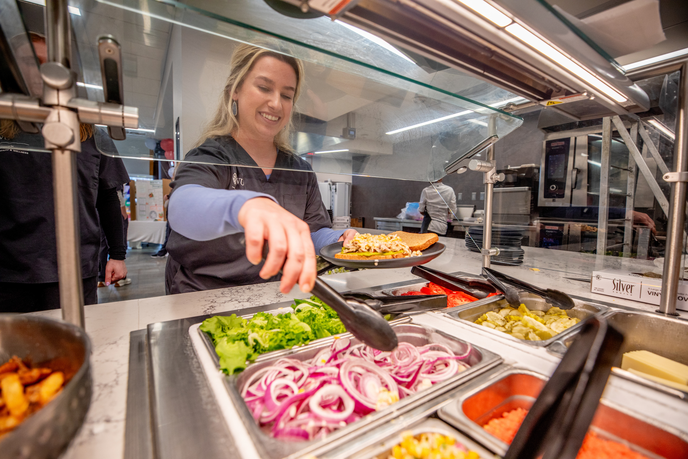 A person smiles while serving a sandwich at a self-serve salad bar with various toppings.