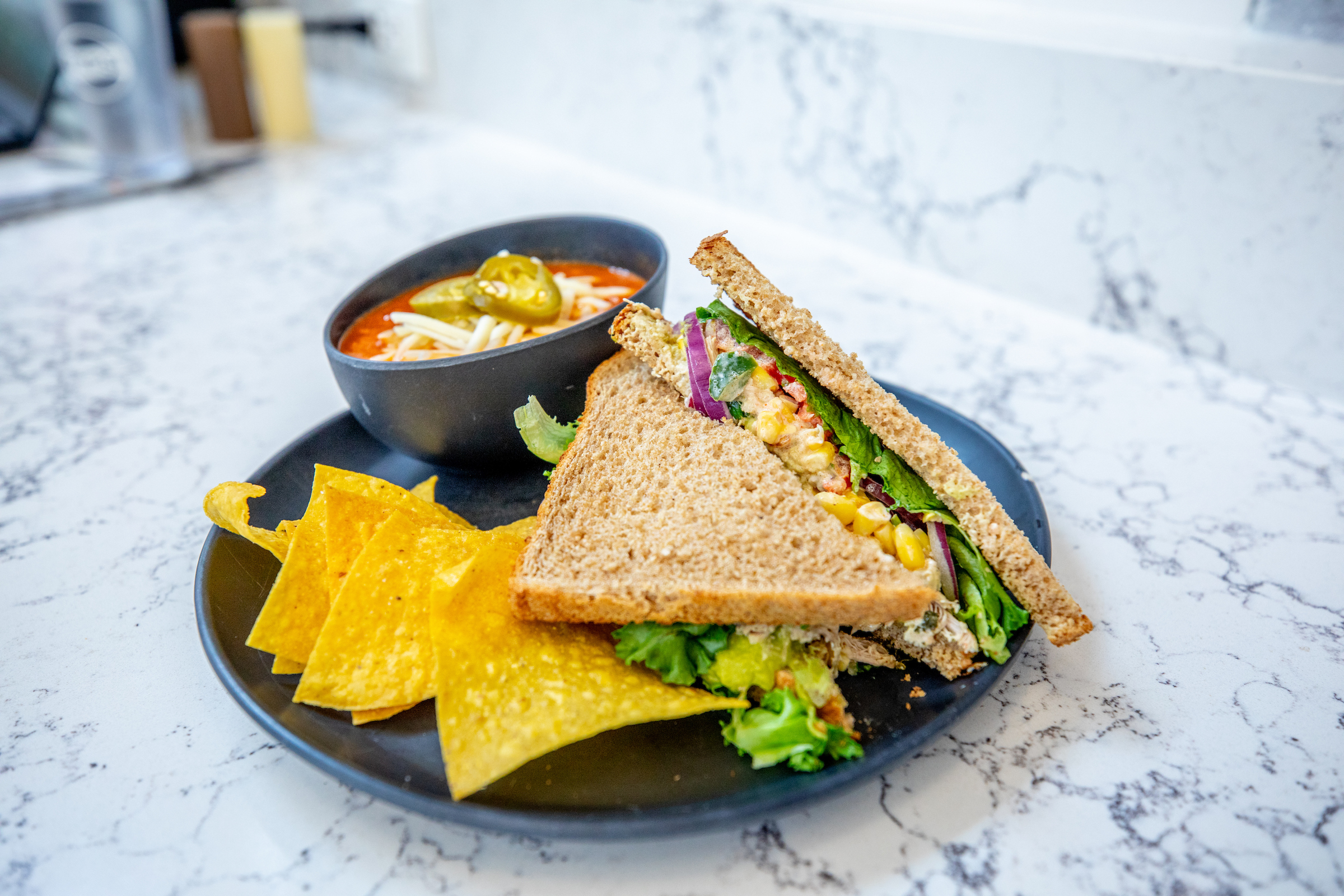 A sandwich with vegetables and corn, served with tortilla chips and a side of soup in a black bowl on a marble countertop.