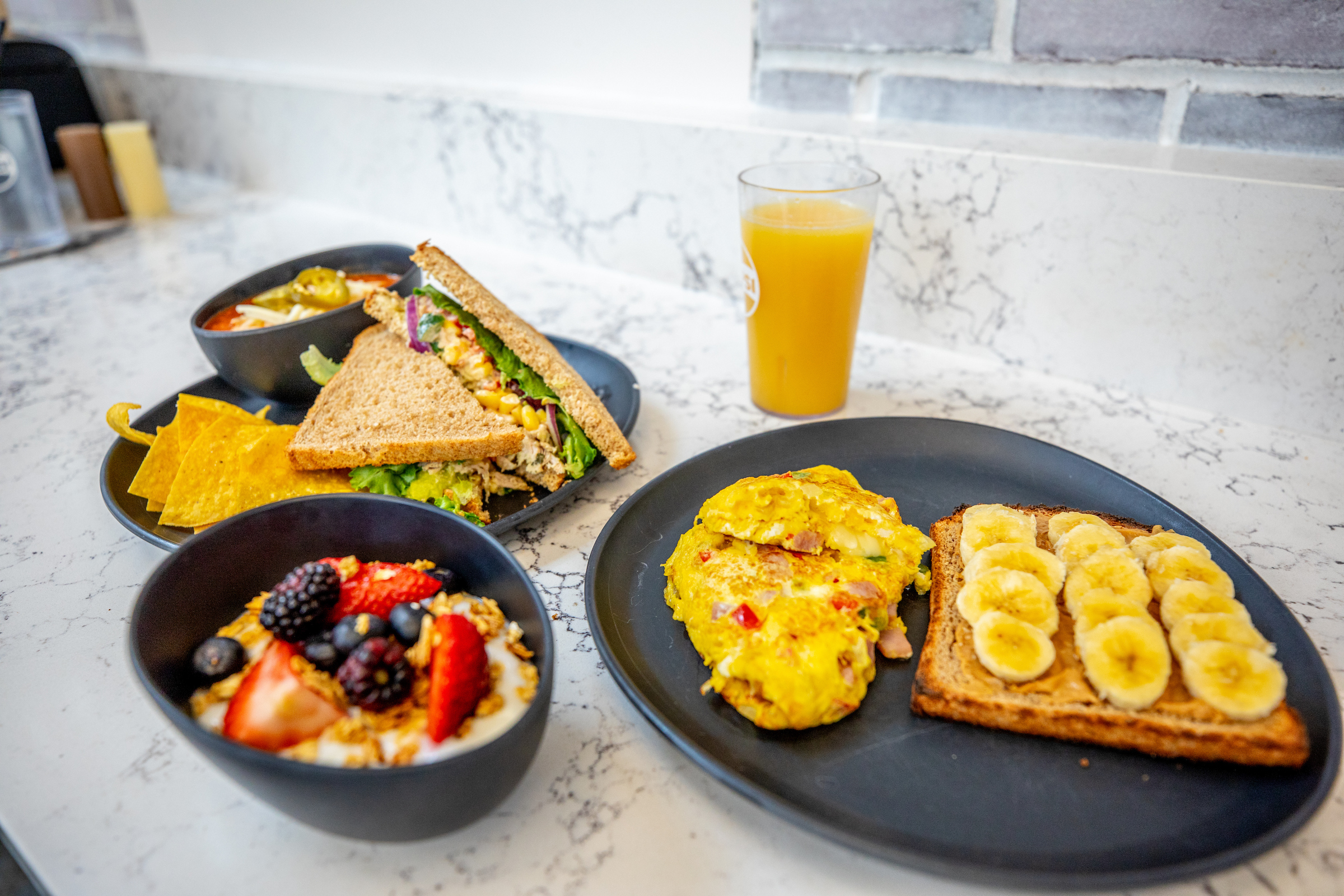 A breakfast spread featuring an omelet with toast topped with banana slices, a fruit and granola bowl, a sandwich, a side of chips, and a glass of orange juice on a marble countertop.