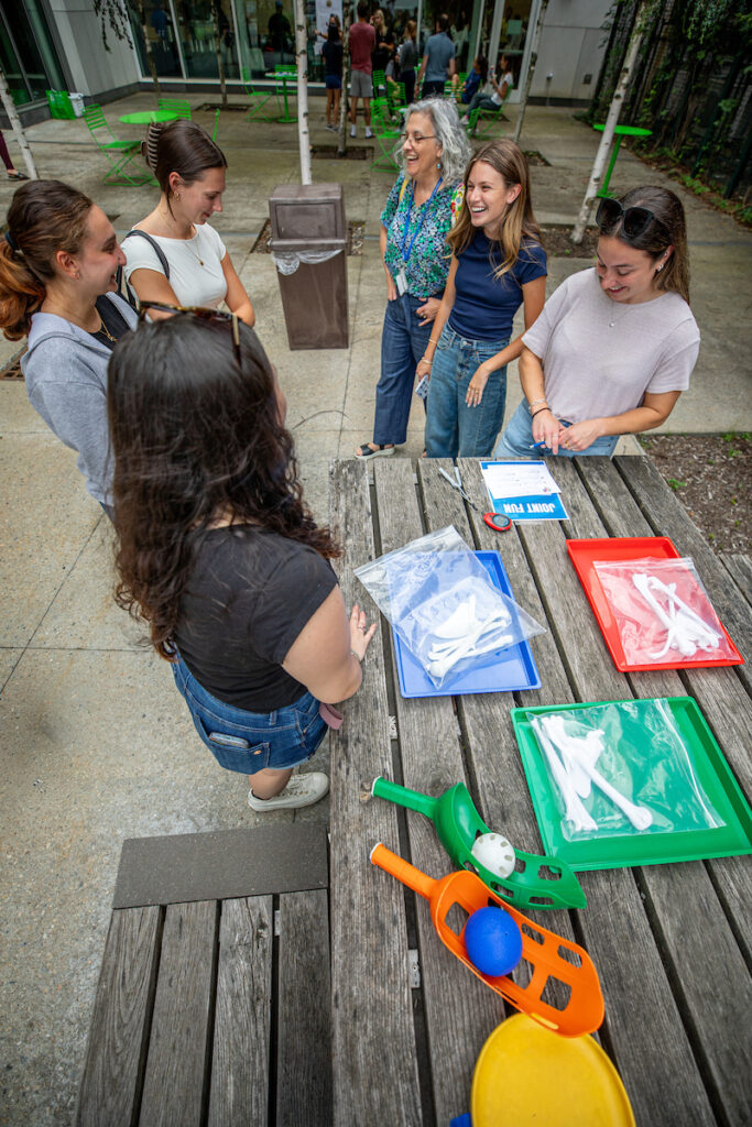 A group of people gather around a wooden table outdoors, engaged in a playful activity involving colorful scoops and balls.