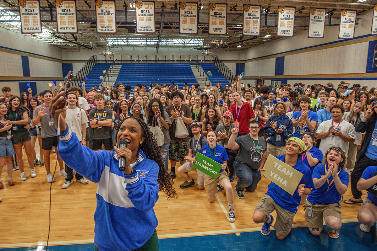 An emcee in a blue and white jacket speaks into a microphone while taking a group selfie on a gymnasium floor, surrounded by a cheering crowd holding signs and celebrating.