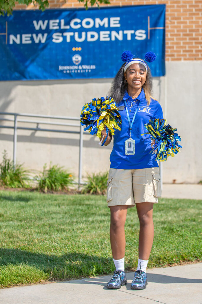 A member of the cat team with pom-poms stands outdoors in front of a Welcome New Students banner.