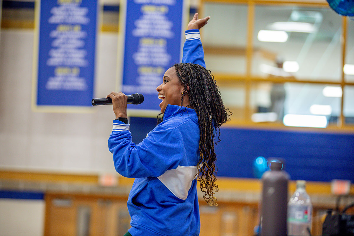An emcee in a blue and white jacket speaks into a microphone while raising one arm in the Wildcat Center.