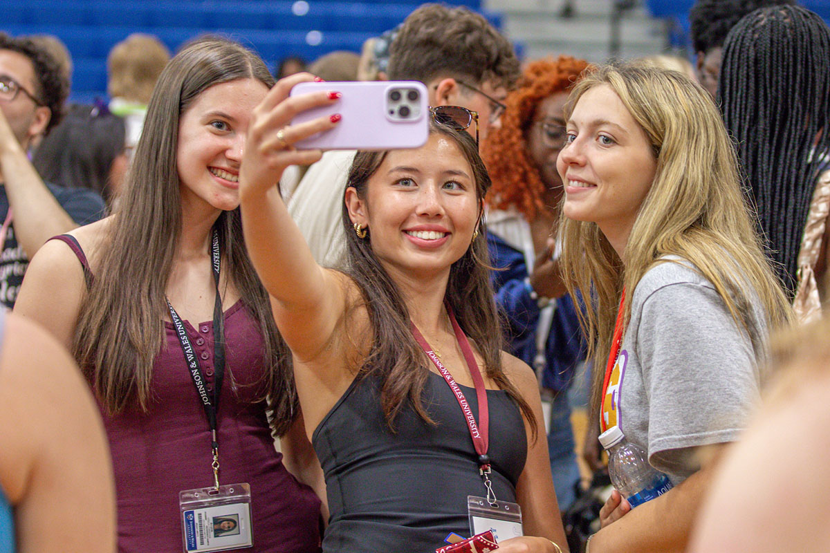 A group of orientation students gather in a gymnasium, with one student holding up a phone to take a selfie with the group.