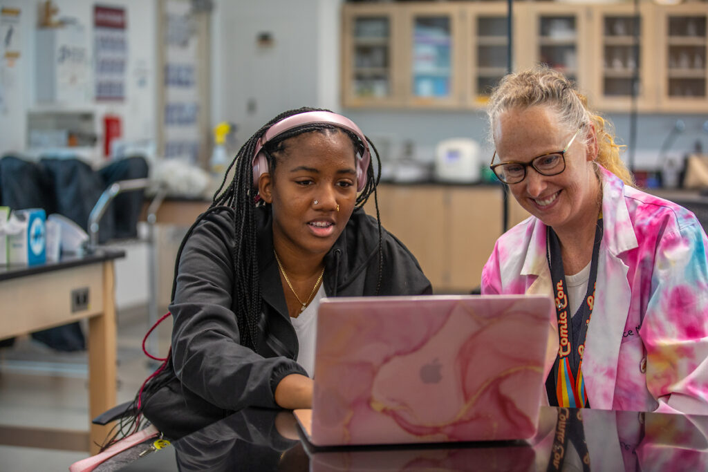 Two individuals collaborate in a lab setting, focused on a laptop with one wearing headphones and the other in a lab coat.