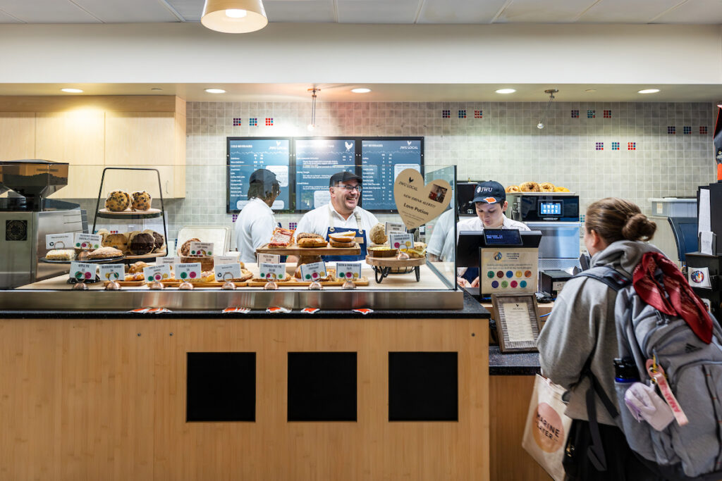 A student stands at the counter of JWU Local, a campus café, while staff members serve sandwiches, pastries and other food items from behind a glass display case. Menu boards and baked goods are visible in the background.