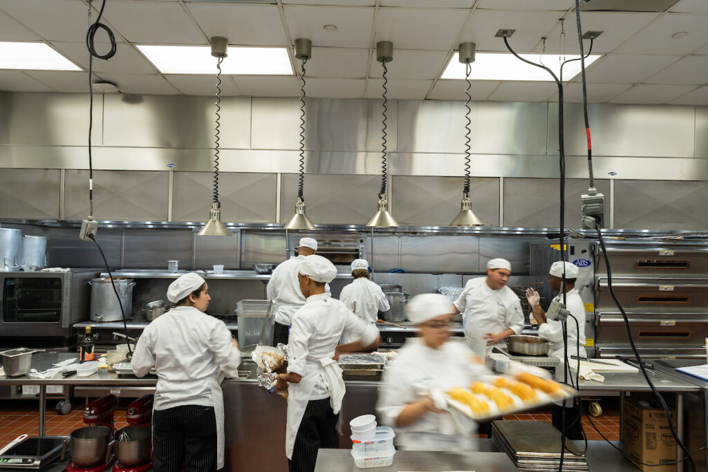 A bustling professional kitchen where chefs in white uniforms work efficiently, preparing food with trays and utensils amidst stainless steel appliances and overhead lighting.