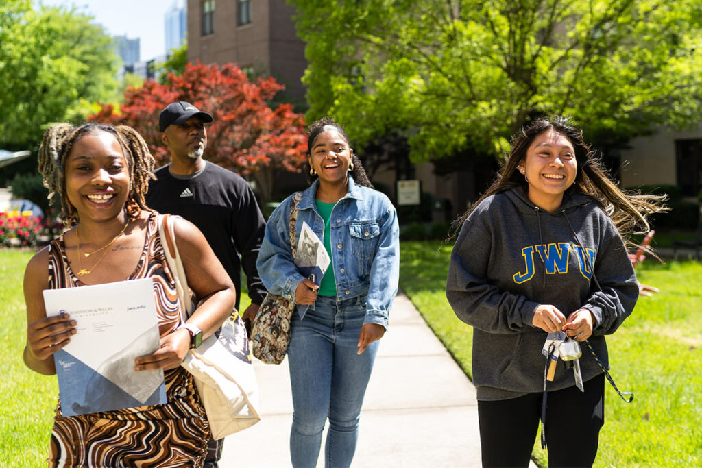 Four students walking through a grassy campus area together on a sunny day.