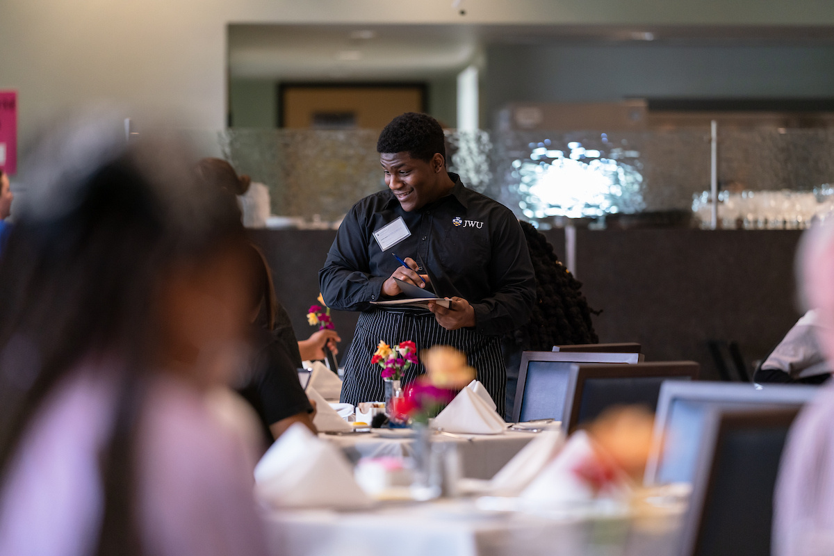 A server wearing a JWU uniform smiles while taking notes at a dining table during an event.