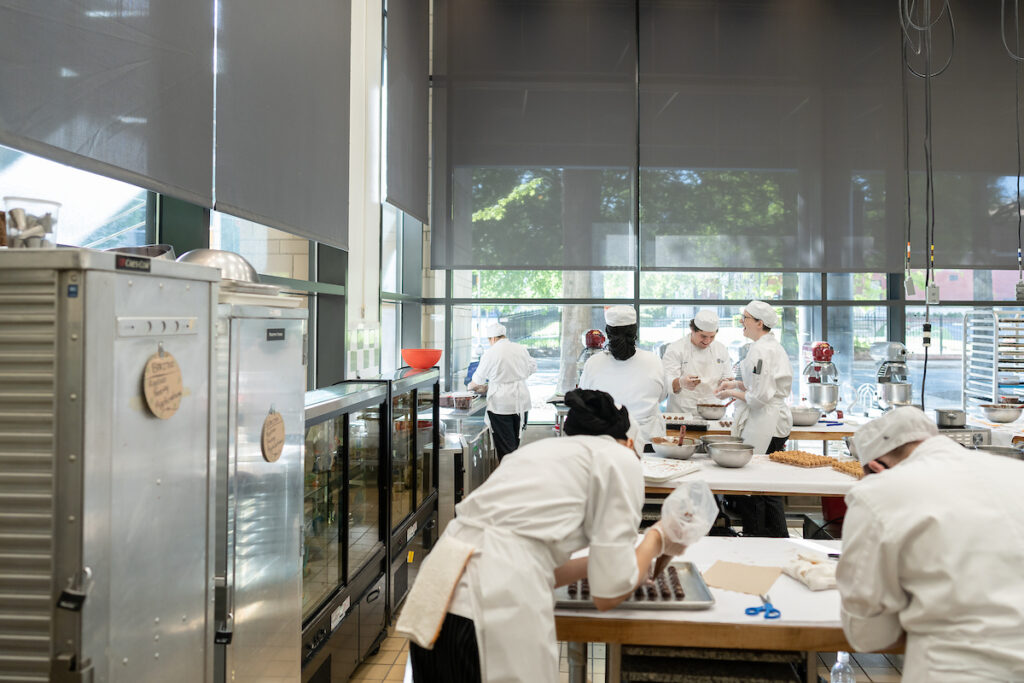 Several individuals in white chef uniforms are working in a professional kitchen, preparing food at stations with large appliances and equipment.