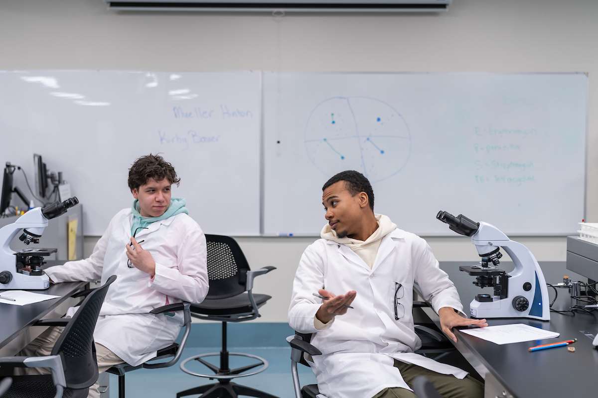 Two individuals in lab coats sit at a desk with microscopes, engaged in discussion in a science classroom.