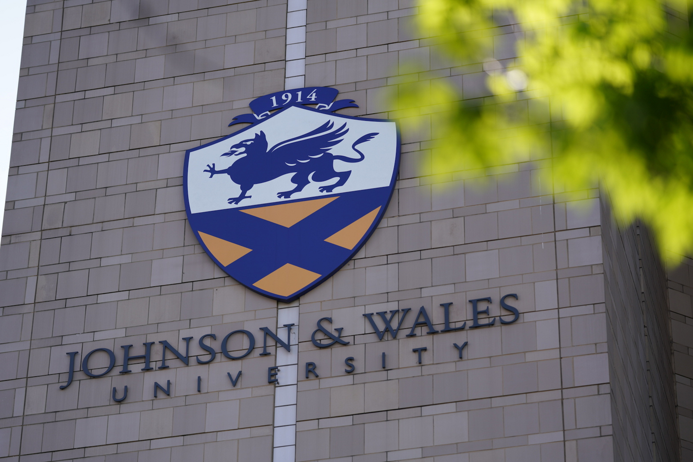 The emblem of Johnson & Wales University, featuring a winged lion and a saltire, is displayed on a stone building facade with green foliage partially obscuring the view.