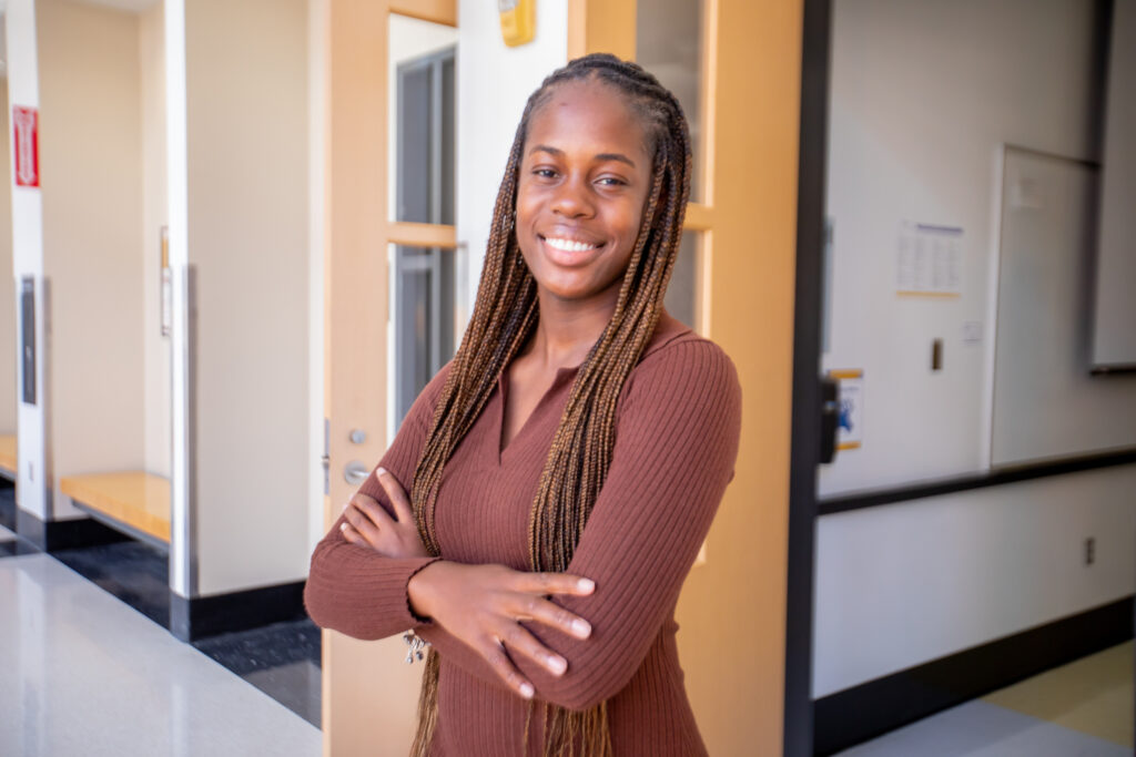 A smiling person with long braided hair stands with arms crossed in a hallway, wearing a brown ribbed top.