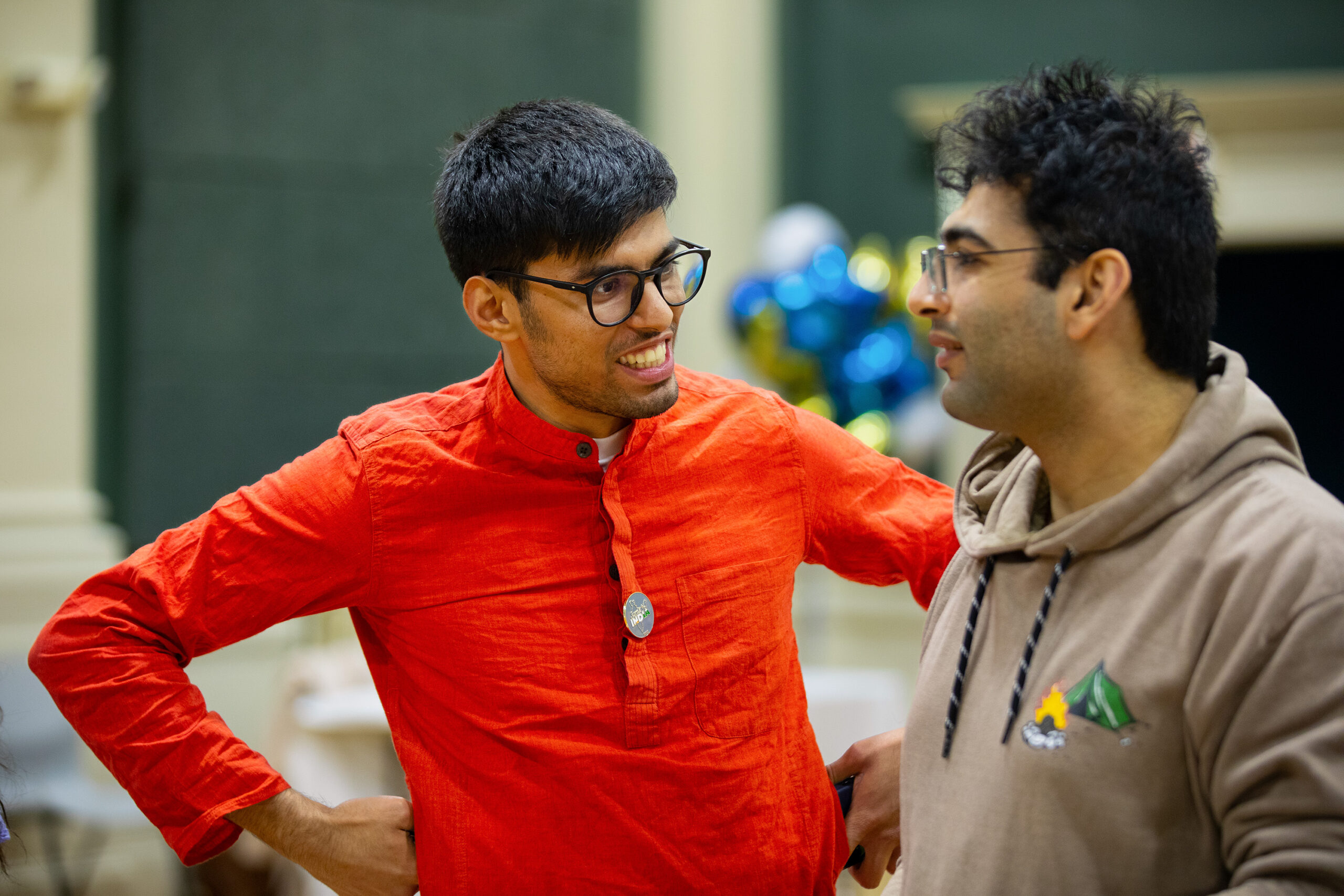 Two individuals engage in lively conversation, one in a vibrant orange shirt and the other in a beige hoodie, with colorful balloons visible in the background.