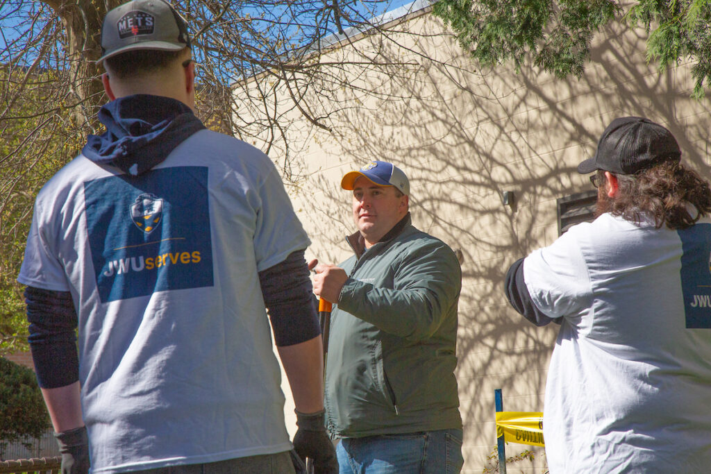 Three individuals wearing "JWU serves" shirts are outdoors, engaged in conversation near a building with tree shadows cast on its wall.