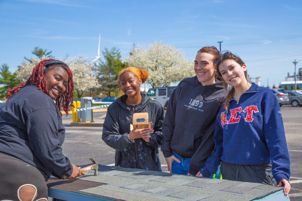 Four young people smile while working on a roof in an outdoor parking lot under a clear blue sky.