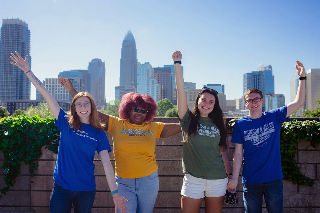 Four students wearing Johnson and Wales University apparel pose joyfully with raised arms against a bright blue sky and the Charlotte skyline.