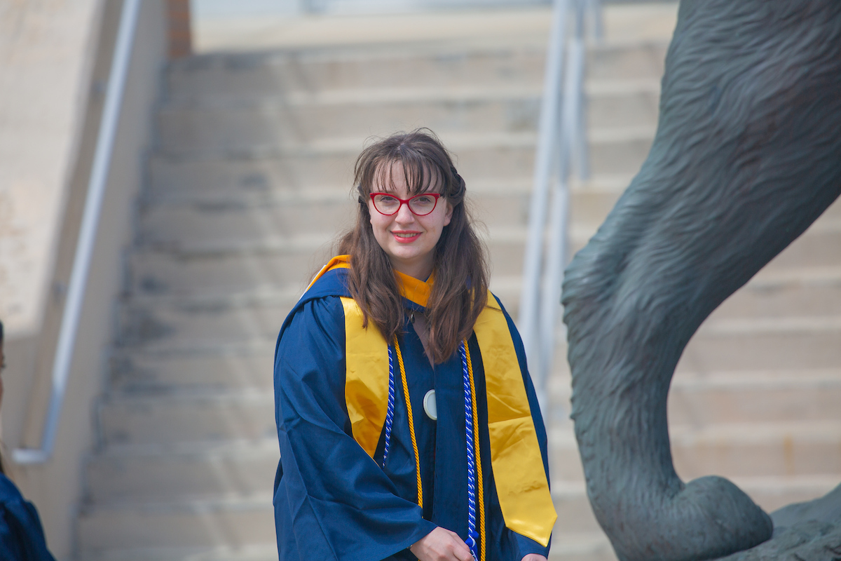 A person in a graduation gown and yellow stole smiles while standing outdoors near stairs and a large sculpture.