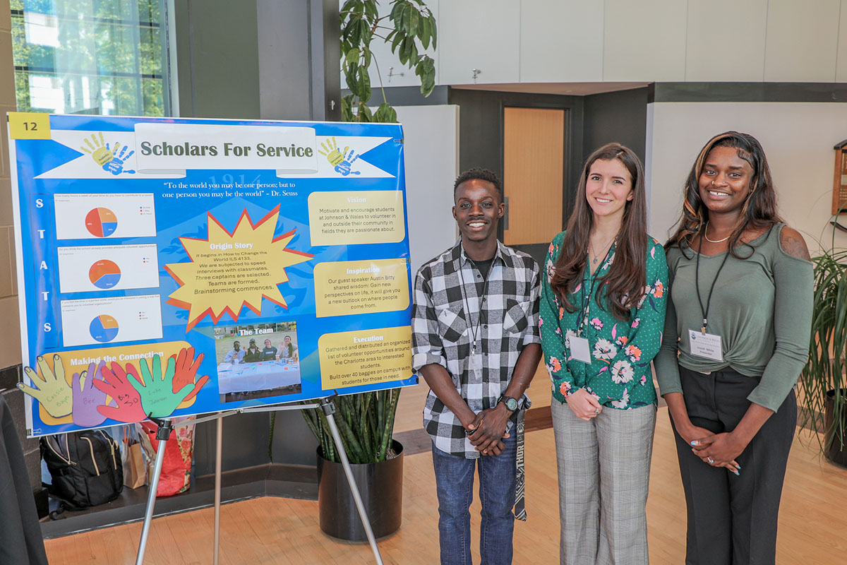 Three students stand beside a colorful display board titled "Scholars For Service" at the Student Research and Design Symposium.