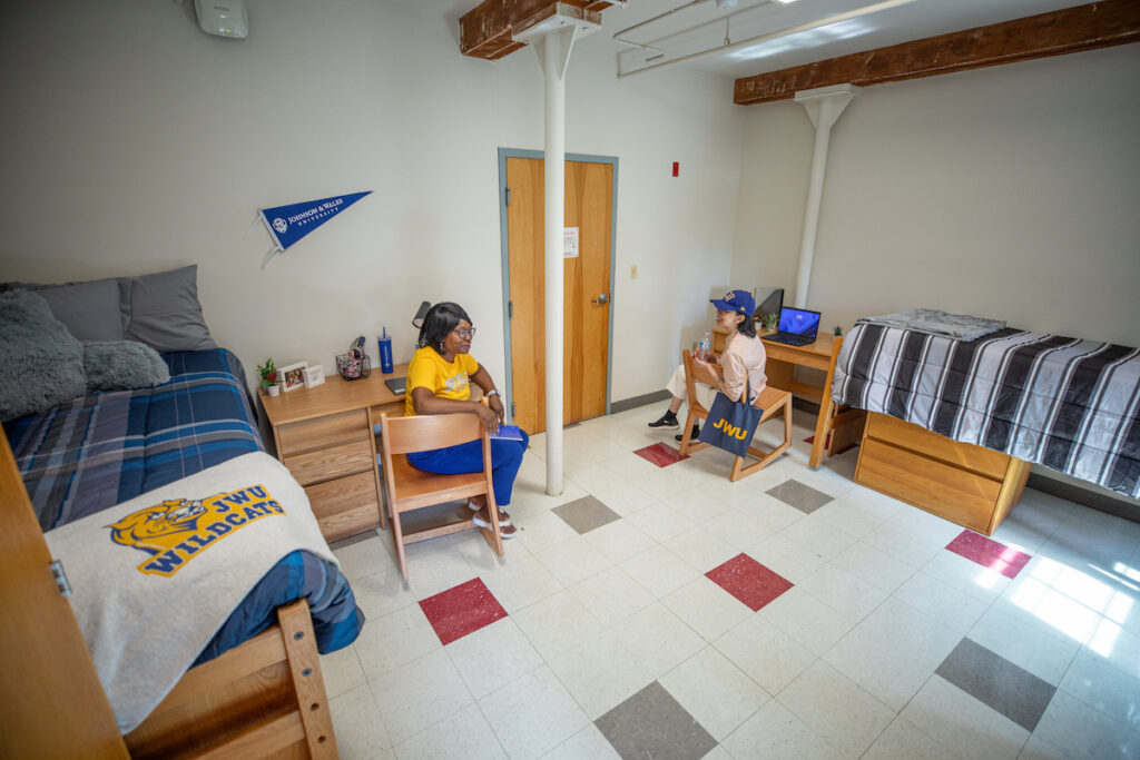 Two individuals sit in a college dorm room with bunk beds, desks, and a flag, engaging in conversation.