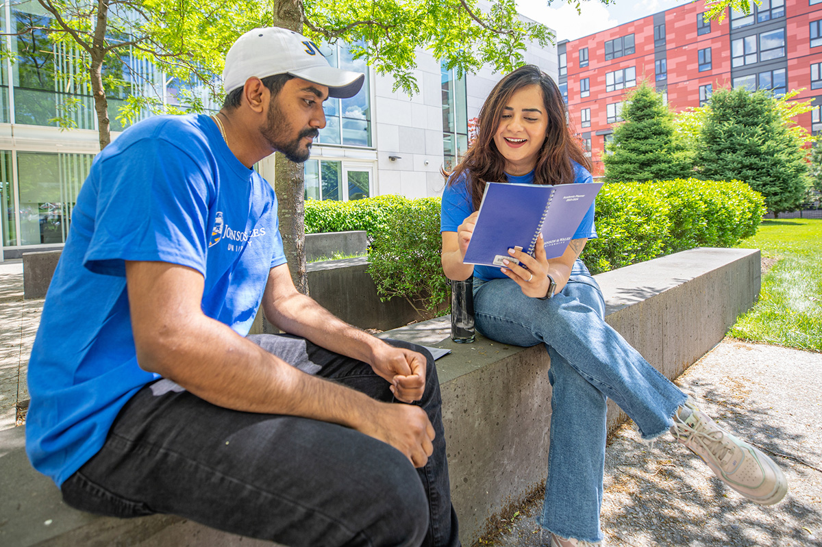 Two individuals sit outdoors on a concrete ledge under trees, engaged in conversation while one holds a notebook.