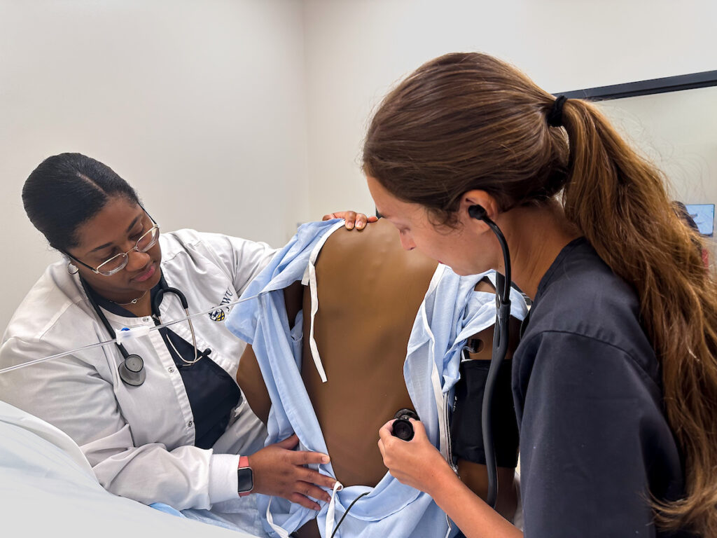 Two medical professionals examine a patient's back using a stethoscope during a clinical training session.