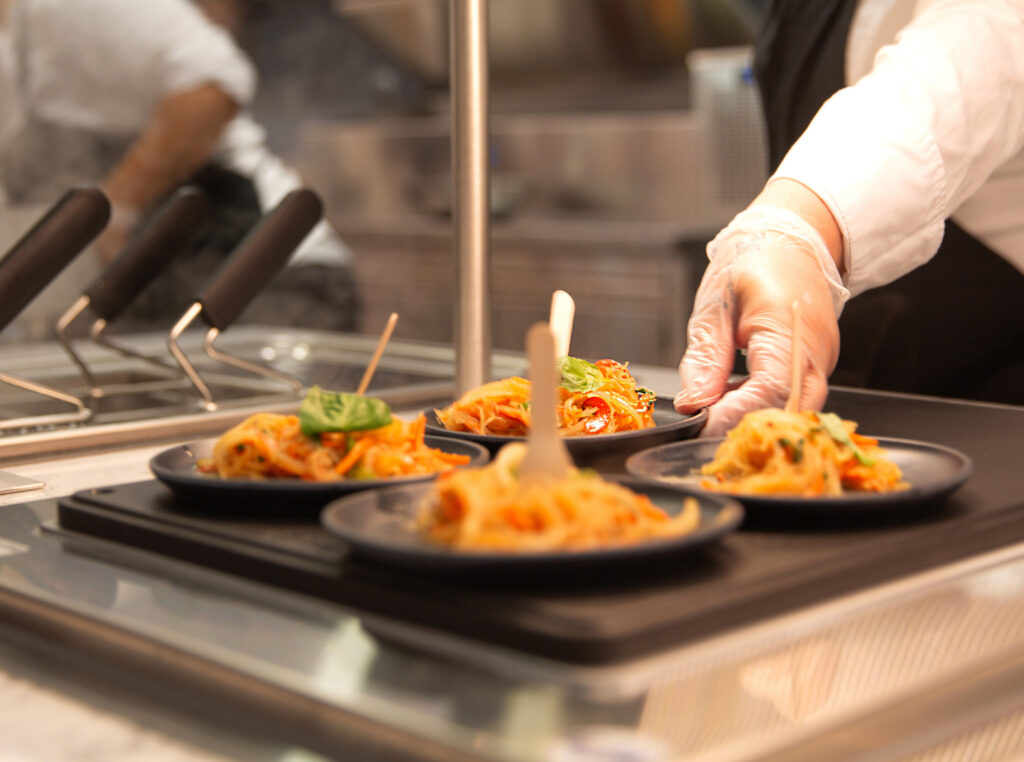 A chef in a professional kitchen arranges plated pasta dishes on a serving station.