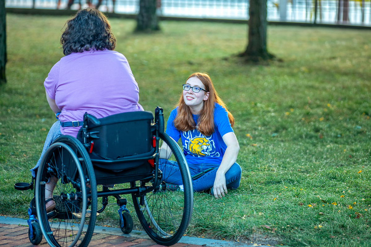 A person in a wheelchair converses with another person sitting on the grass in Gaebe Commons.