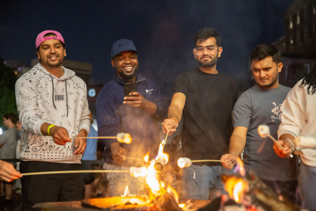 A group of students roast marshmallows over a fire at night.