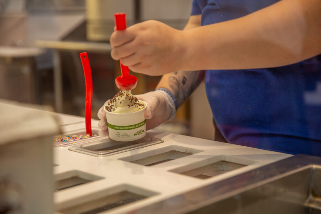A person in a blue shirt and gloves uses a red spoon to sprinkle chocolate shavings onto a cup of ice cream at a frozen yogurt station.