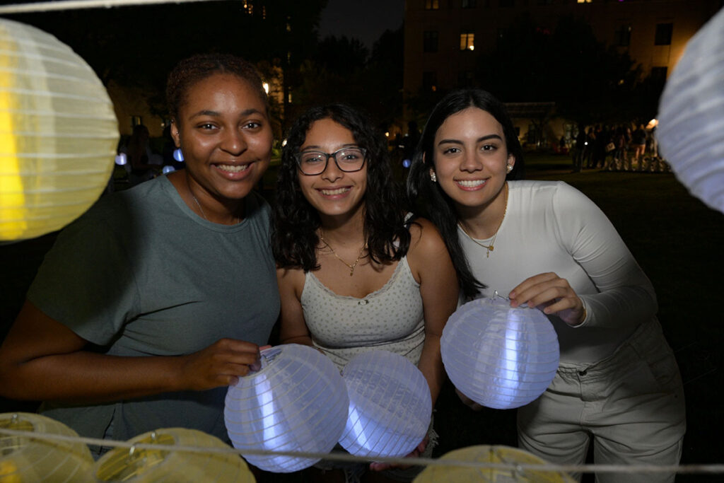 Three students smile while holding illuminated paper lanterns outdoors at night.