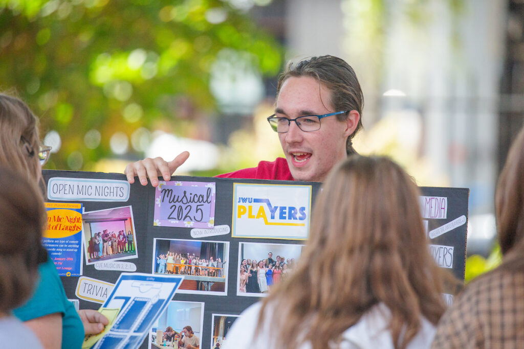 A person wearing glasses points to a promotional board advertising campus events like open mic nights, a musical, and trivia, while engaging with attendees outdoors.