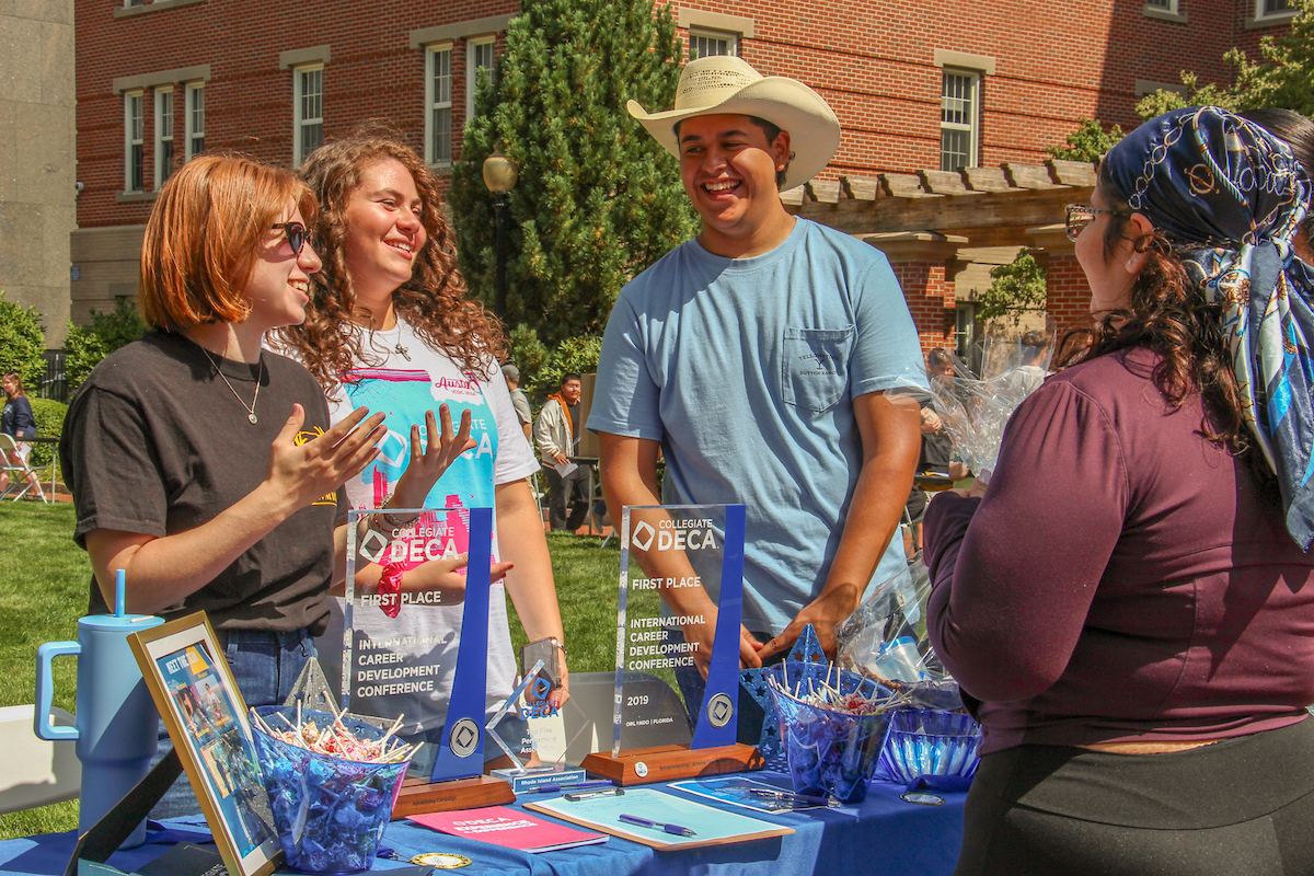 Four individuals celebrate on a sunny day outdoors, standing behind a table displaying “First Place” awards from the Collegiate DECA International Career Development Conference 2019.