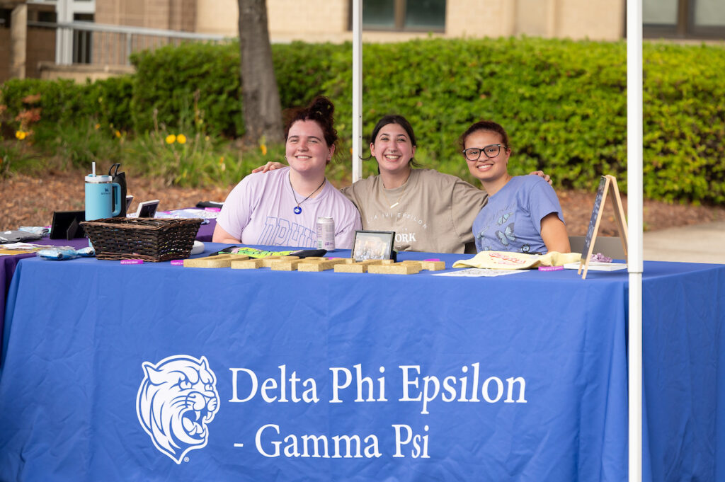 Three smiling individuals stand behind a blue table covered with a Delta Phi Epsilon - Gamma Psi banner, promoting their sorority at an outdoor event.