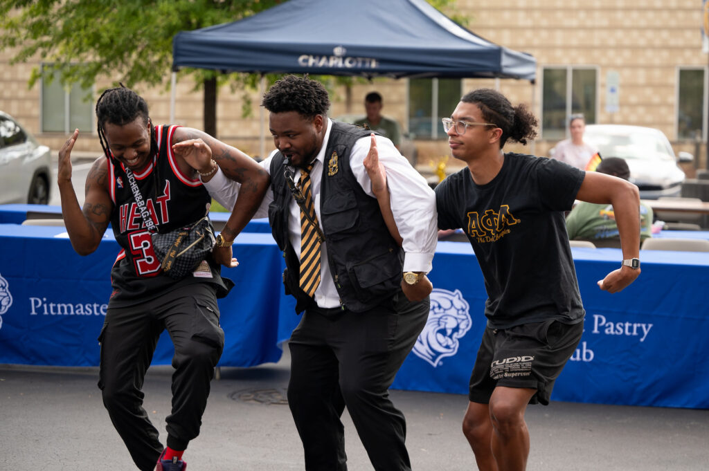 Three individuals are dancing energetically outdoors, with one in a basketball jersey, another in a vest and tie, and the third in a black t-shirt and shorts, in front of blue event banners.