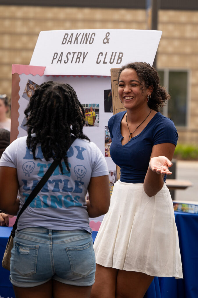 A person in a blue top and white skirt smiles while offering something to another person wearing a t-shirt that reads "Enjoy the Little Things," standing in front of a "Baking & Pastry Club" display.