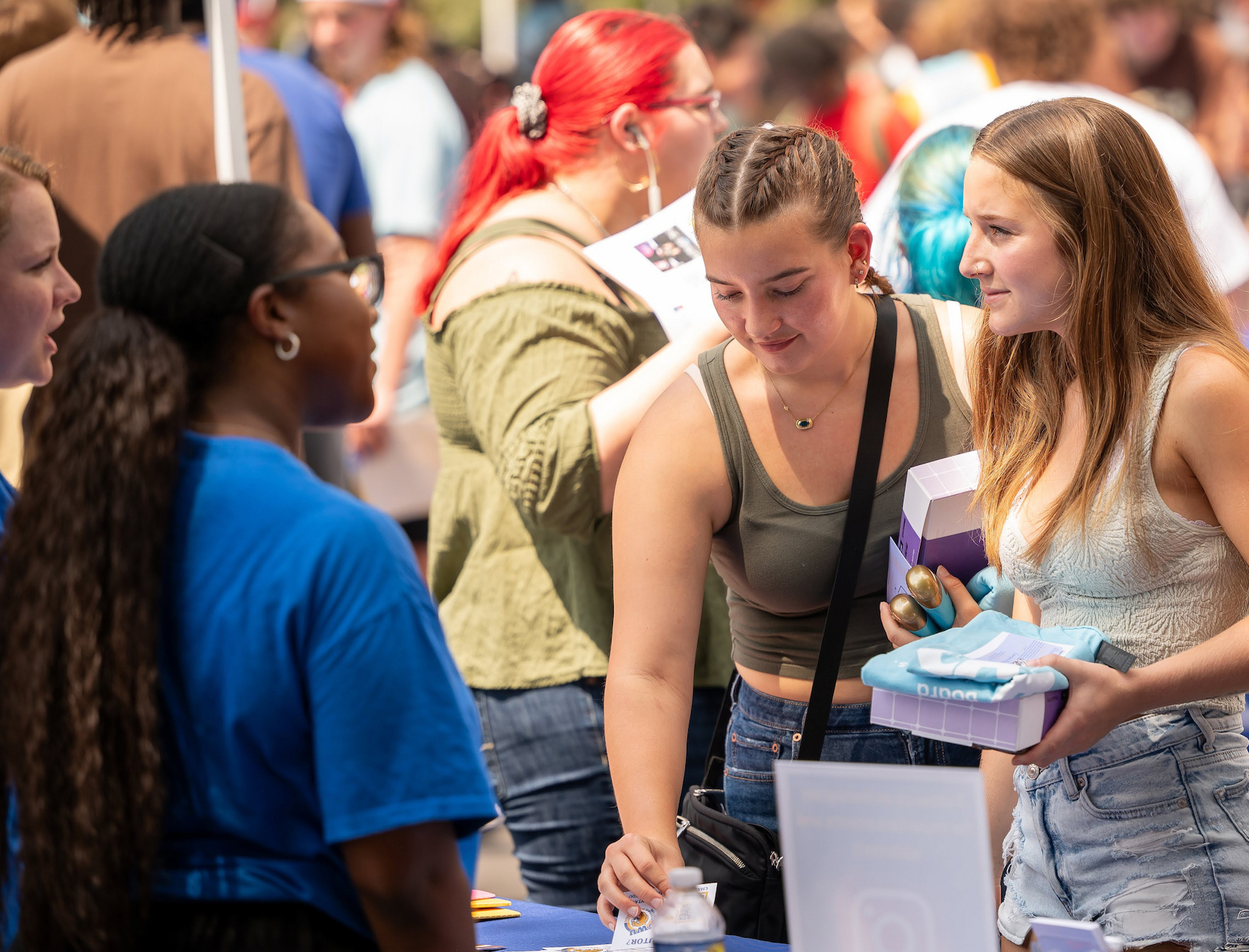 Two individuals interact at an outdoor event, examining items on a table while others gather around under tents.
