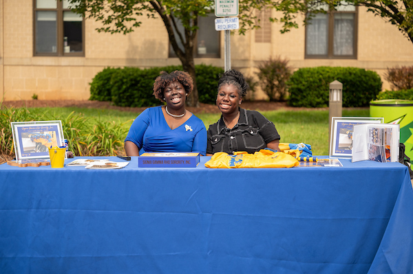 Two individuals smile warmly behind a blue table displaying promotional materials and a yellow garment for the Sigma Gamma Rho Sorority, Inc.