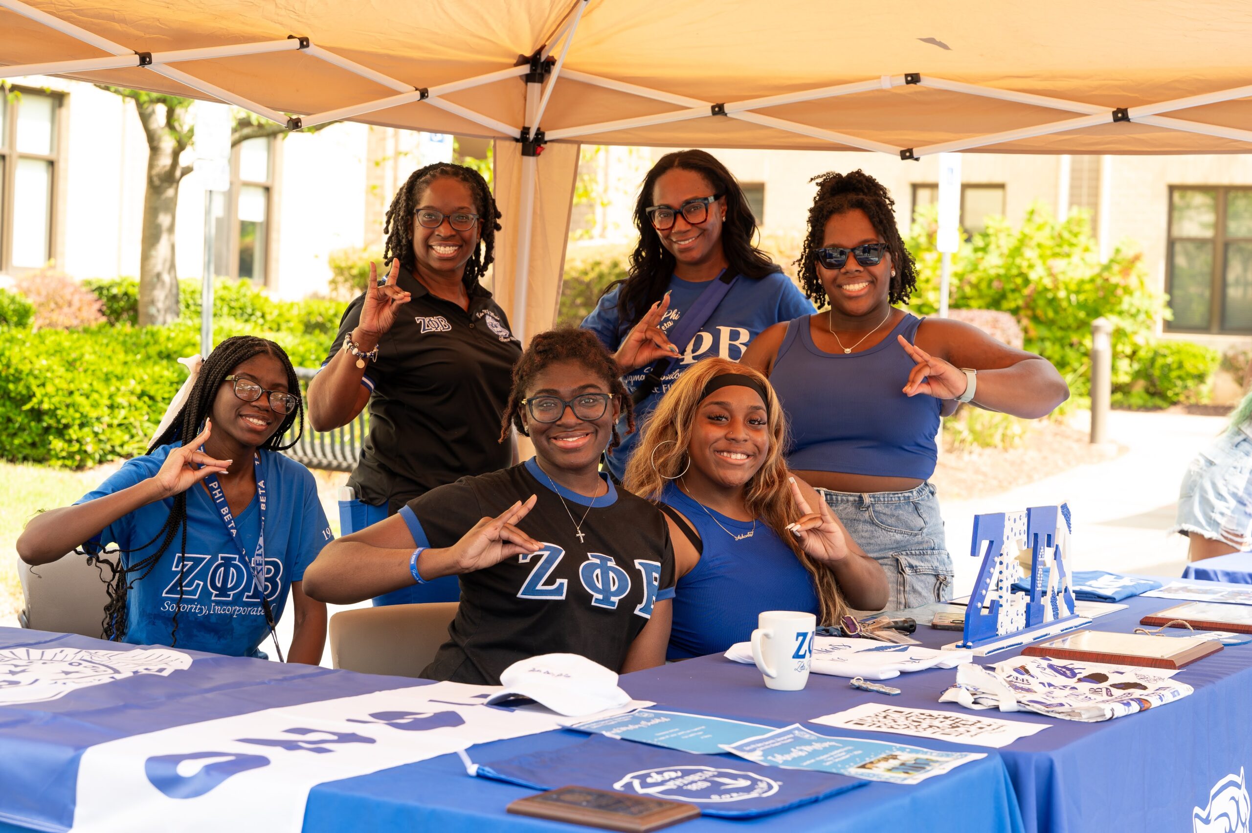 A group of six individuals in Zeta Phi Beta sorority attire pose cheerfully at an outdoor booth, making hand gestures and smiling for the camera.