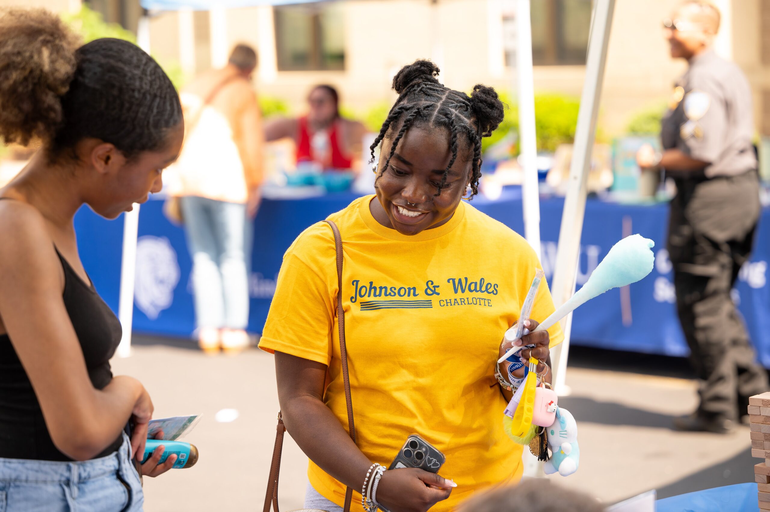 A person in a yellow Johnson & Wales Charlotte shirt smiles while holding a cotton candy stick and keychain at an outdoor event.