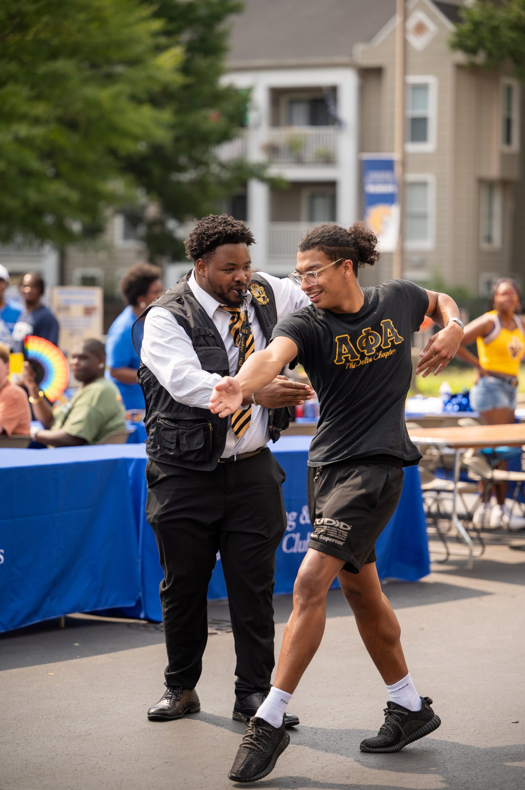 Two individuals engage in a lively interaction outdoors, one wearing a vest and tie while the other sports a black t-shirt with Greek letters.