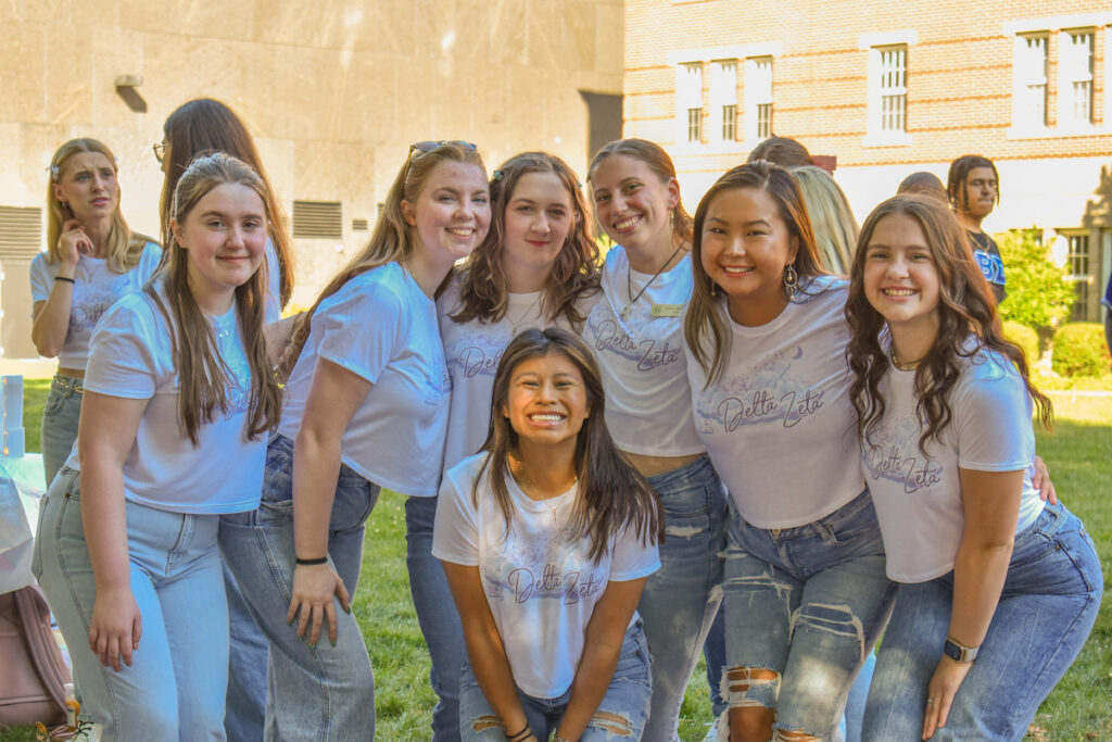 A group of young people wearing "Delta Zeta" shirts pose together outdoors in front of a brick building.