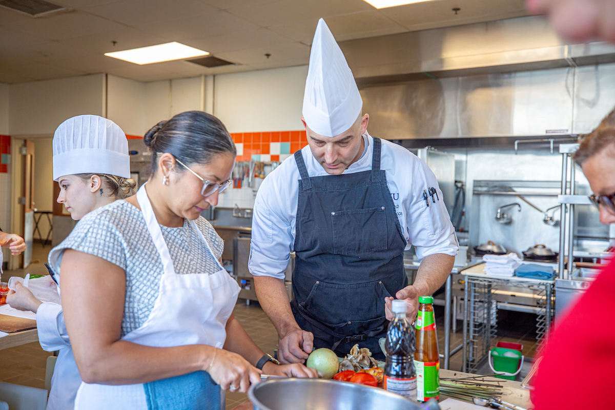 A chef in a tall white hat and black apron demonstrates a cooking technique to participants in a professional kitchen setting.