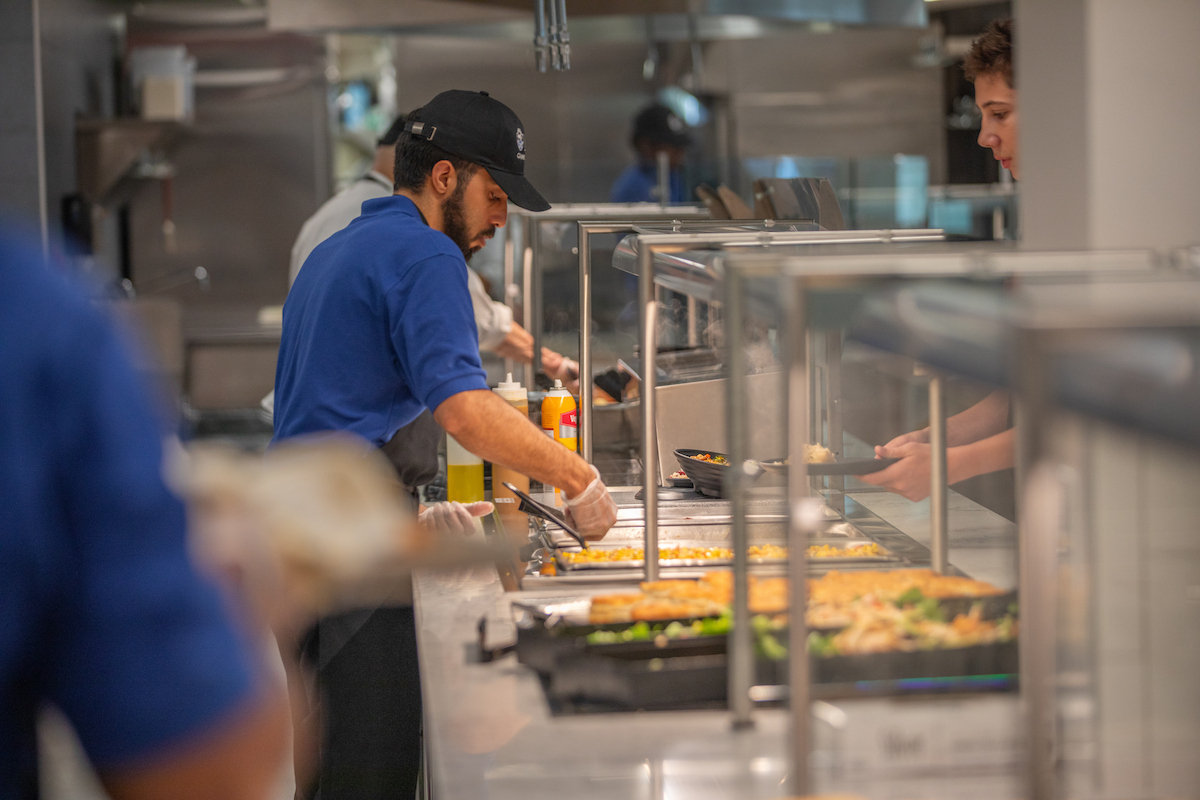 A person in a blue shirt and black cap serves food at a cafeteria counter while another individual receives a plate.