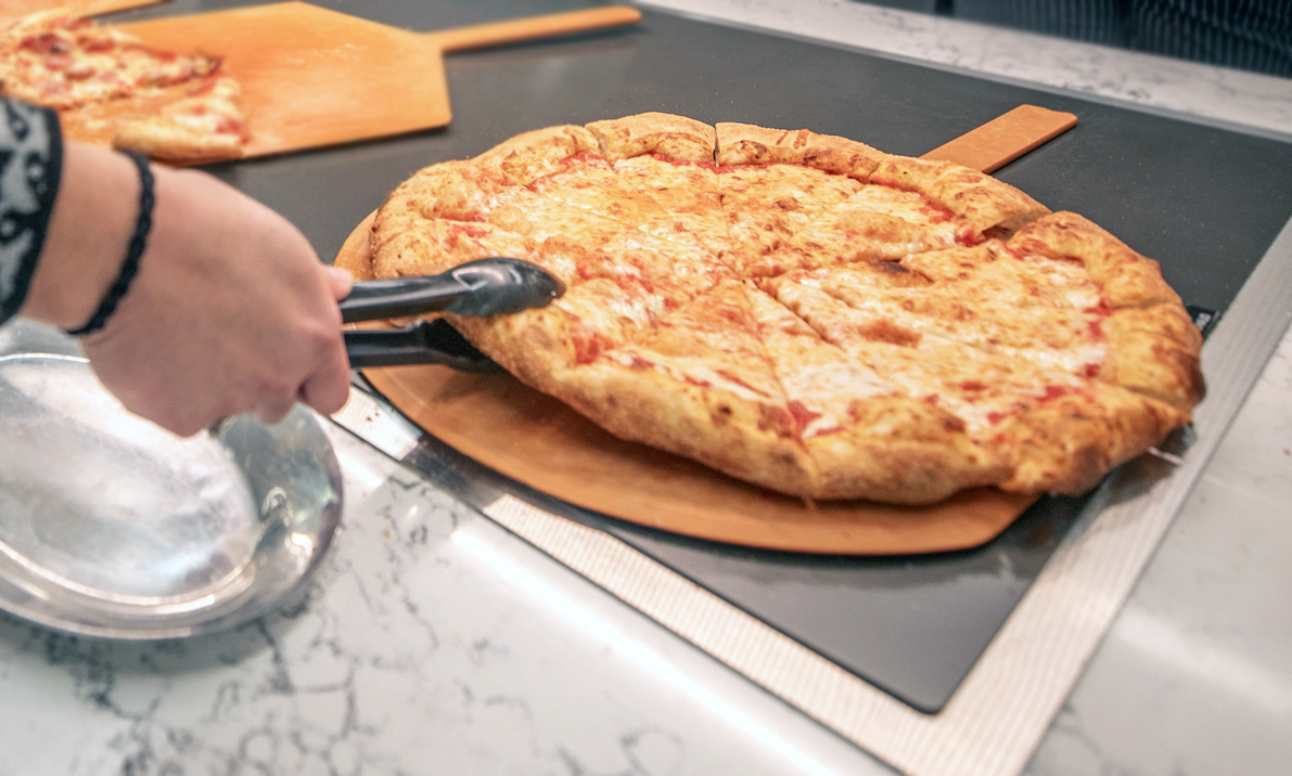 A person selects a slice of pizza from a buffet counter in a modern kitchen setting.