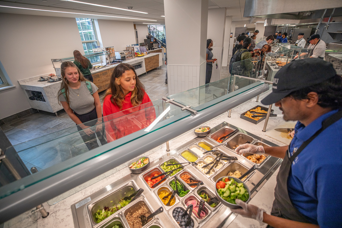A person in a blue shirt and black cap serves food from a salad bar to two individuals standing at the counter.