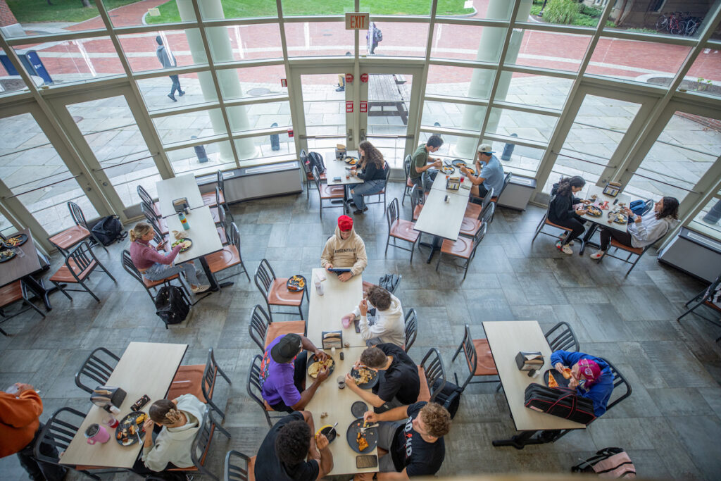 Students eat lunch in a spacious, well-lit cafeteria with large windows overlooking an outdoor area.