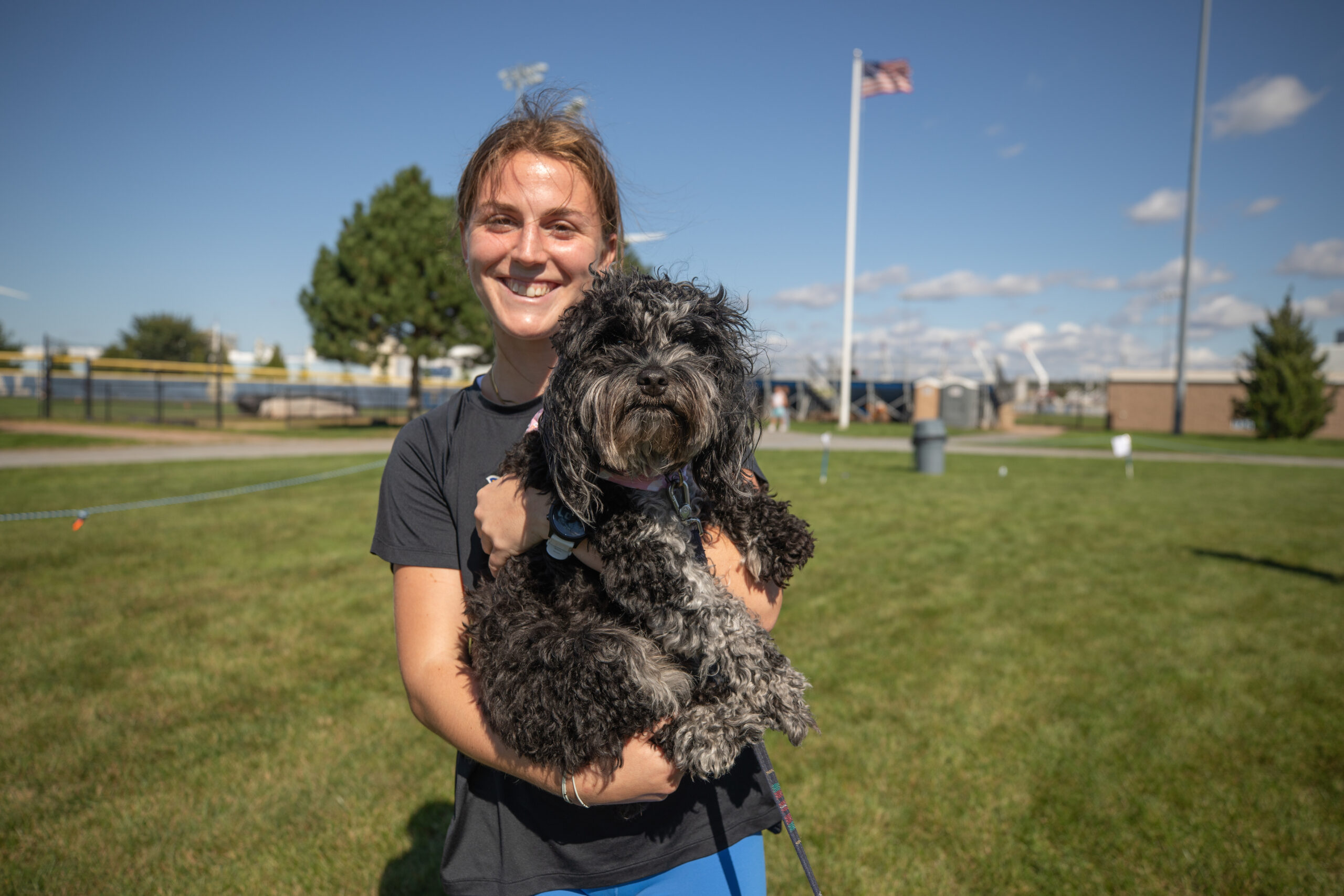 A person smiles while holding a black and gray dog outdoors on a sunny day.